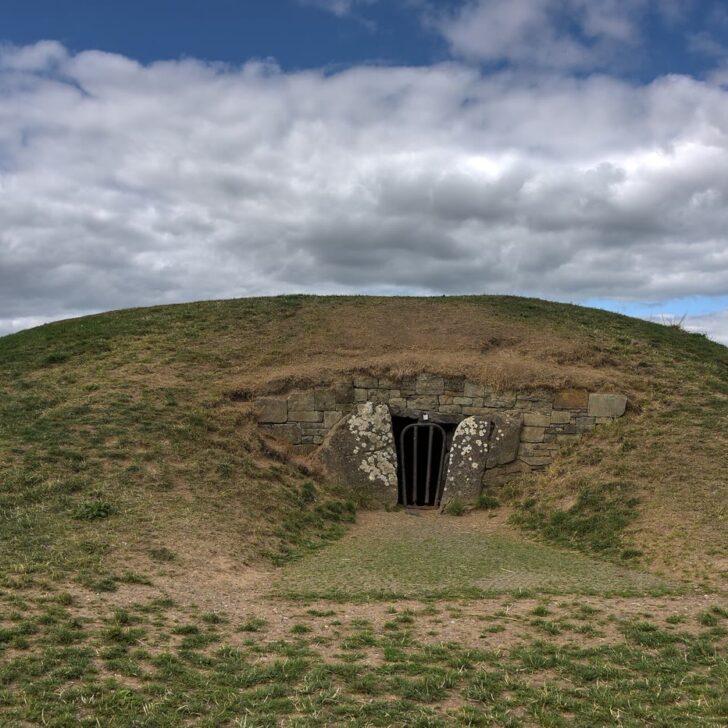The Mound of the Hostages on the Hill of Tara: a grassy Neolithic passage tomb mound with a small barred stone entrance under a big cloudy sky