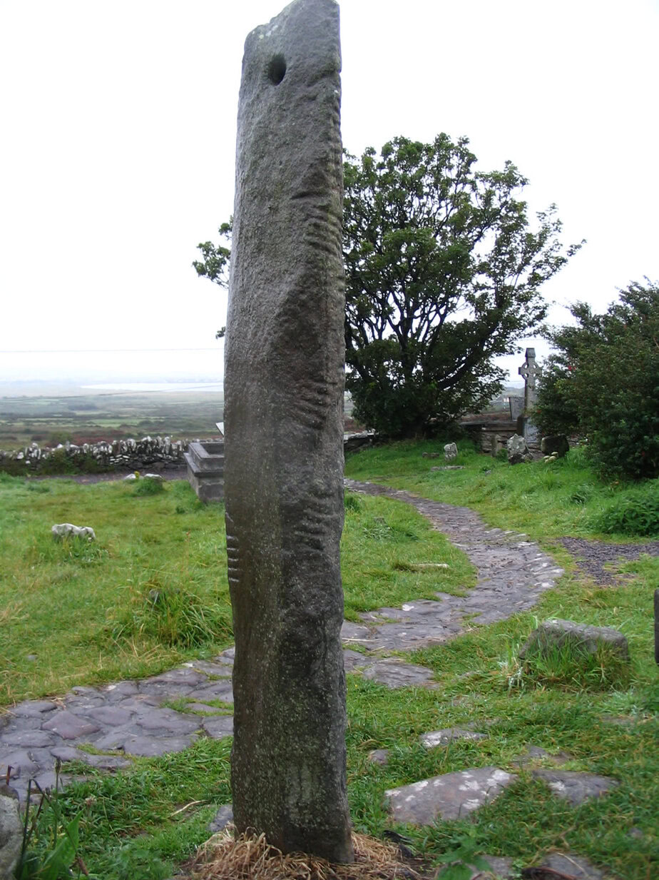 Kilmalkedar ogham stone: a tall, narrow grey pillar with a round hole near the top and grooved ogham strokes along one edge, standing in a grassy churchyard in Co. Kerry