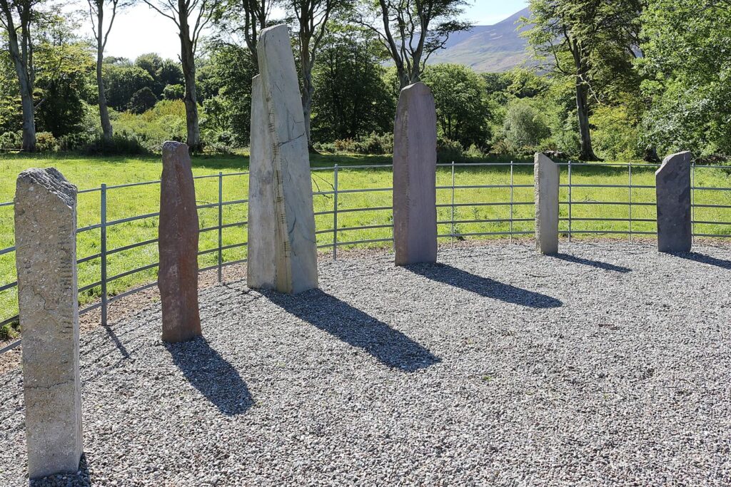 Dunloe Ogham Stones: tall standing stones with ogham strokes cut along their edges, set in a curve on gravel in front of a metal railing near the Gap of Dunloe, Co. Kerry, with trees and a mountain behind