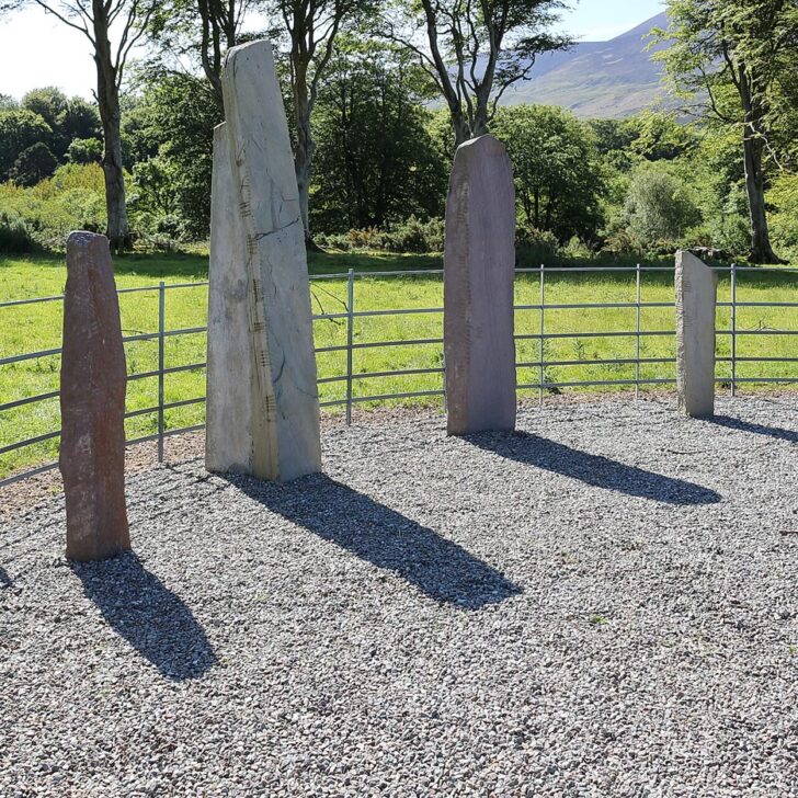 Dunloe Ogham Stones: tall standing stones with ogham strokes cut along their edges, set in a curve on gravel in front of a metal railing near the Gap of Dunloe, Co. Kerry, with trees and a mountain behind