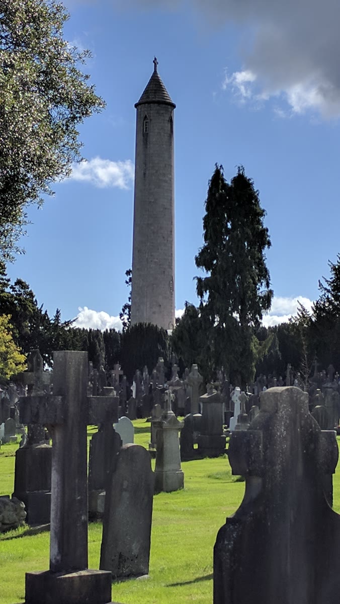 The O'Connell Tower, a tall round tower with a conical cap topped by a cross, rising above old gravestones and dark trees in Glasnevin Cemetery, Dublin