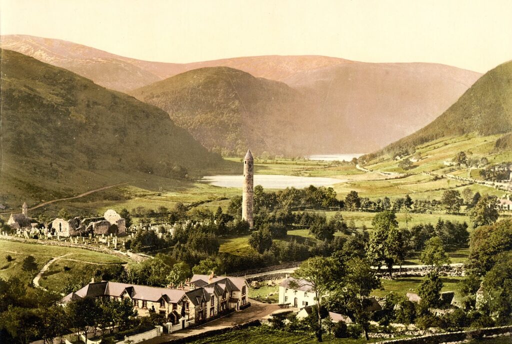 Irish round towers: 1890s colour photochrom of the round tower at Glendalough rising above trees in a green Wicklow valley, with monastic ruins to the left and the lake and hills behind