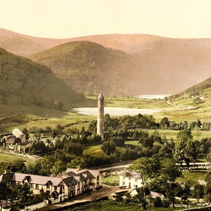 Irish round towers: 1890s colour photochrom of the round tower at Glendalough rising above trees in a green Wicklow valley, with monastic ruins to the left and the lake and hills behind