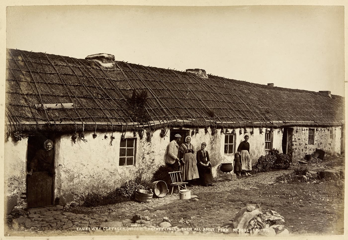 1880s photograph of a long thatched cottage near the Giant's Causeway, with an old woman in a shawl at one door and a small group of people standing outside