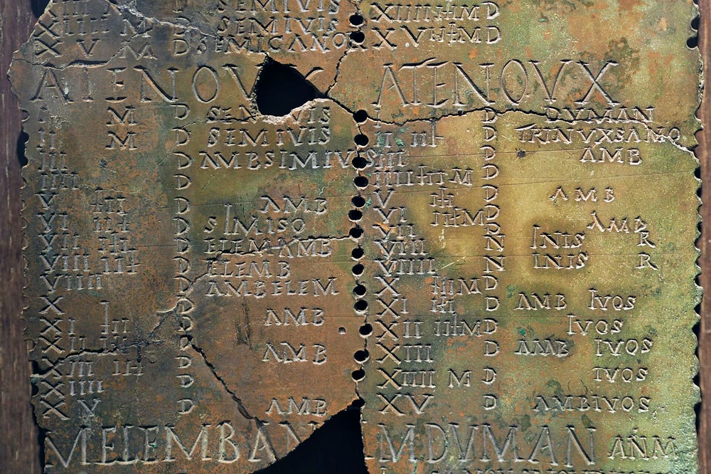 Close-up of the Gaulish Coligny calendar: a cracked green-brown bronze tablet inscribed with Roman capitals in columns, with a line of small peg holes running down the middle