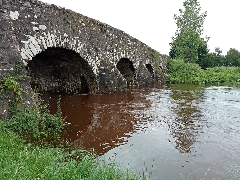 The River Moyola running high and peat-brown under a many-arched old stone bridge, with long grass on the bank and trees beyond