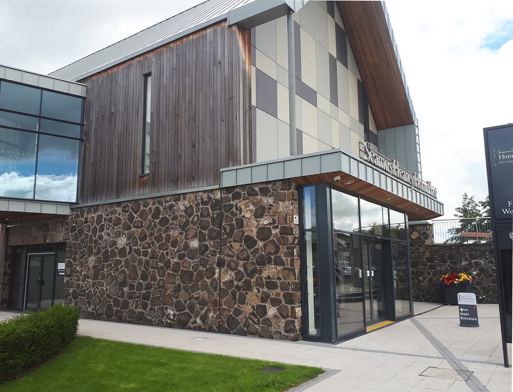 The entrance of Seamus Heaney HomePlace in Bellaghy: a modern building with dark timber cladding above rough dark stone walls, and a glass doorway under a sign bearing the centre's name