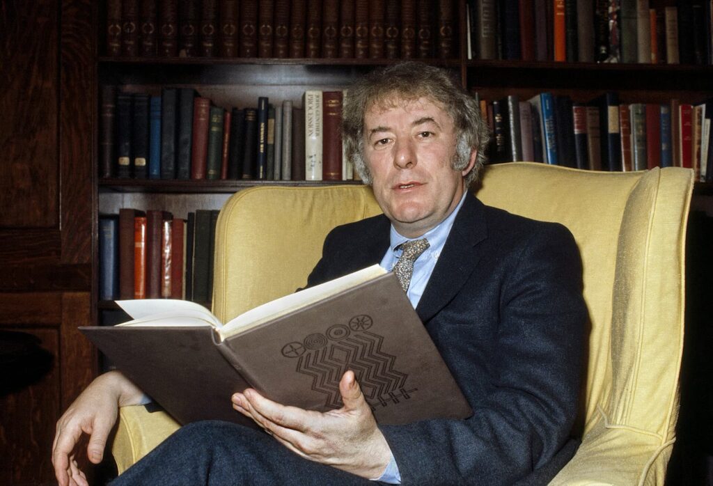 Seamus Heaney in 1982, with grey curly hair, a dark jacket and tie, sitting in a yellow wing armchair in front of tall bookshelves and holding an open book