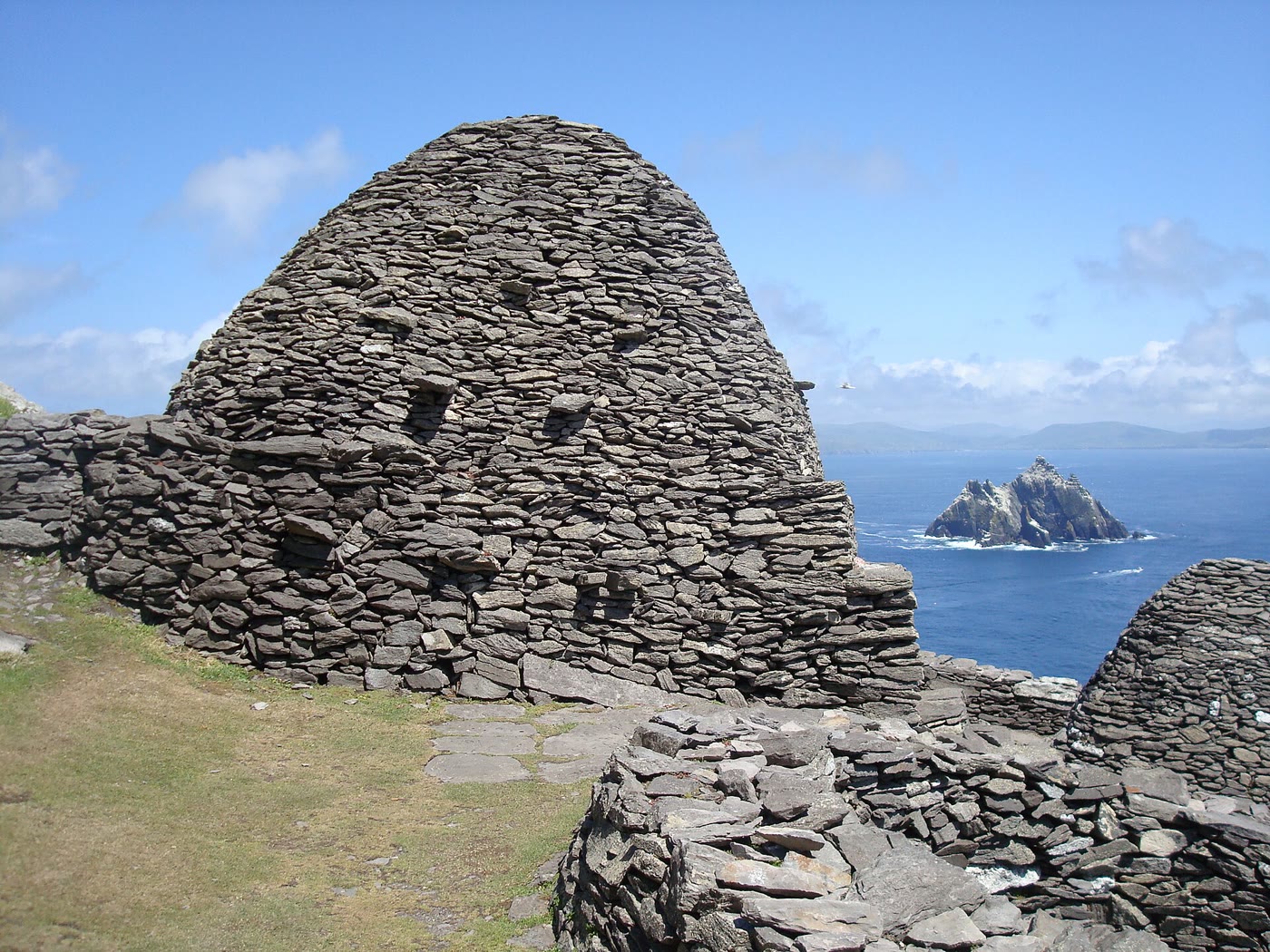 Skellig Michael beehive huts: a tall dome of dry-stone slabs beside lower stone walls, with the jagged rock of Little Skellig, streaked white with gannets, in a blue sea behind