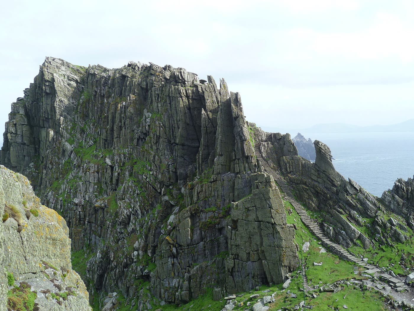 A long flight of stone steps climbing a narrow green ridge beside towering grey sandstone cliffs on Skellig Michael, with Little Skellig faint on the sea behind