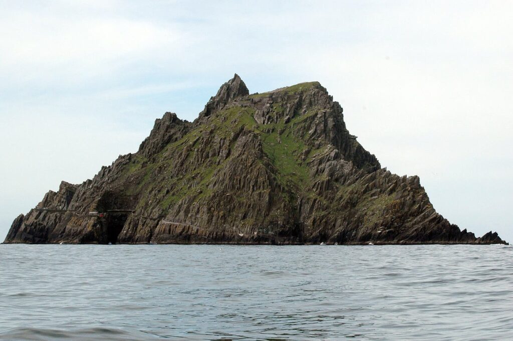 Skellig Michael rising steeply from a calm grey sea: a jagged green and dark sandstone island with twin peaks, under a pale sky