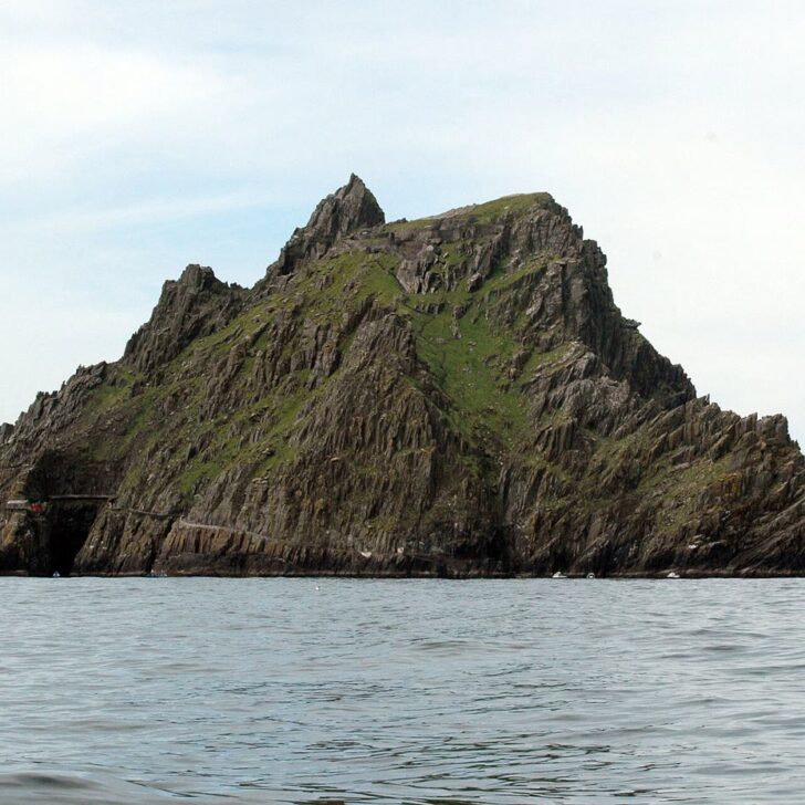 Skellig Michael rising steeply from a calm grey sea: a jagged green and dark sandstone island with twin peaks, under a pale sky