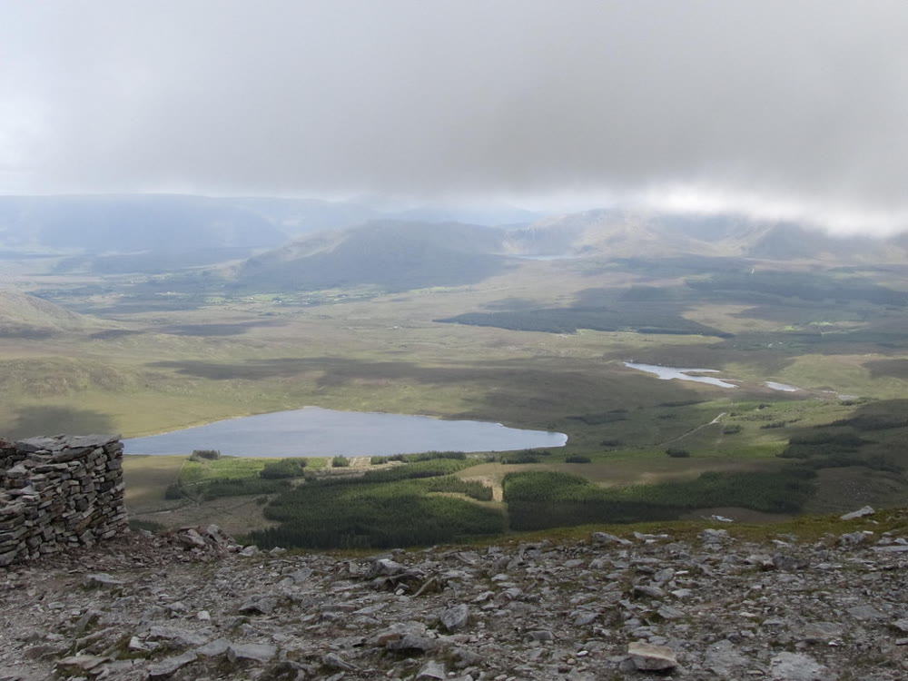 View from the stony summit of Croagh Patrick down to Lough Nacorra, a long grey lake among bog and forestry, with hills under low cloud beyond