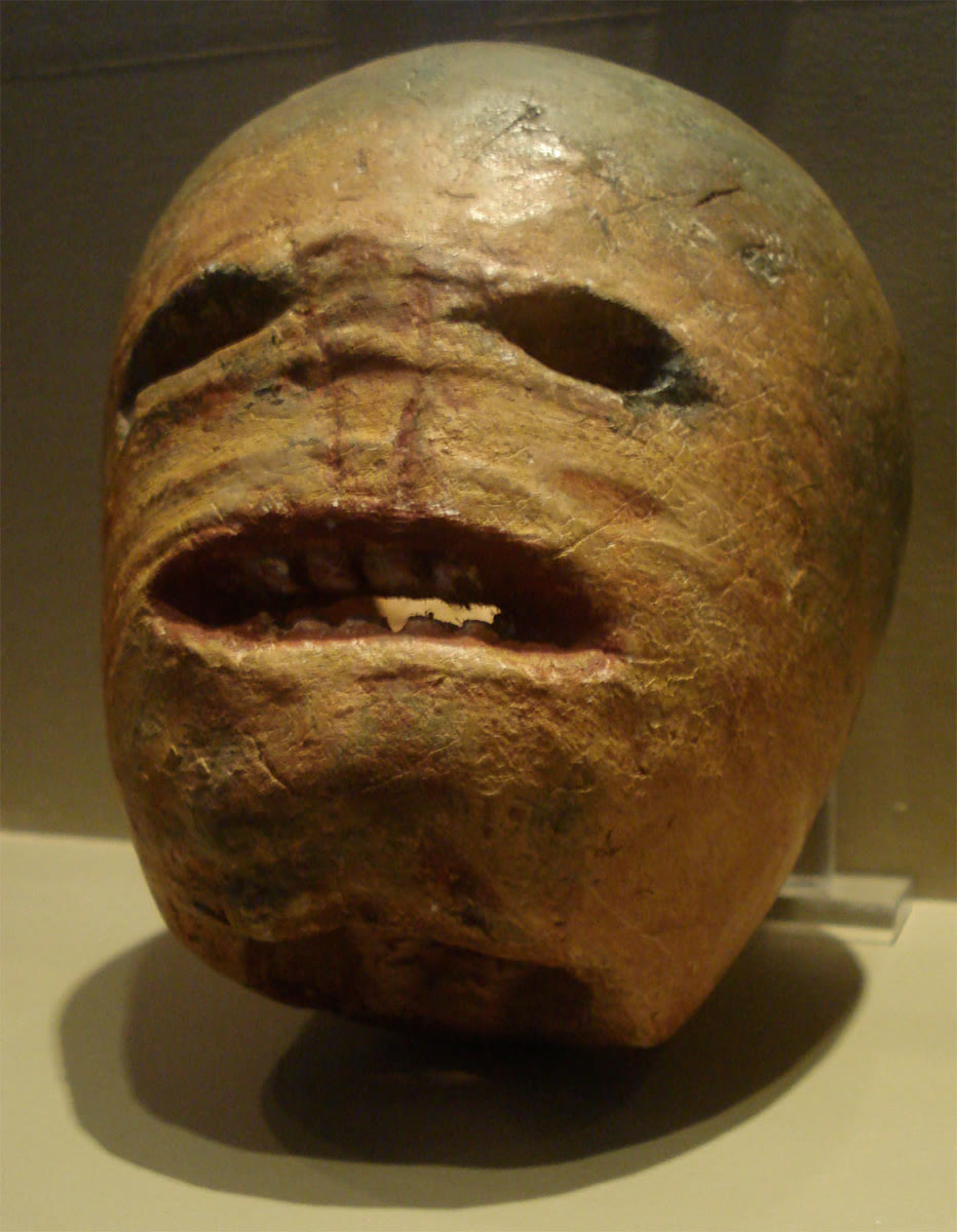 Close-up of a shrivelled, carved turnip lantern with hollow eye holes, a gaping mouth and crooked teeth, on a stand in a museum case