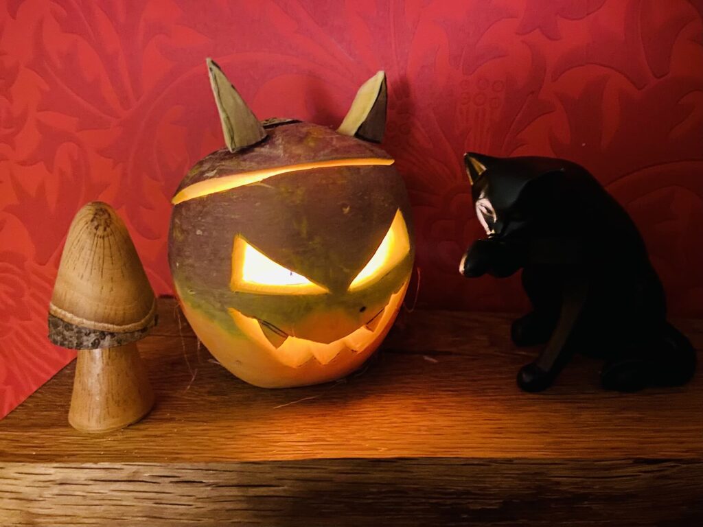 A carved turnip lantern with slanted glowing eyes, a jagged grin and pointed ears, lit from inside, beside a small wooden toadstool and a black cat ornament