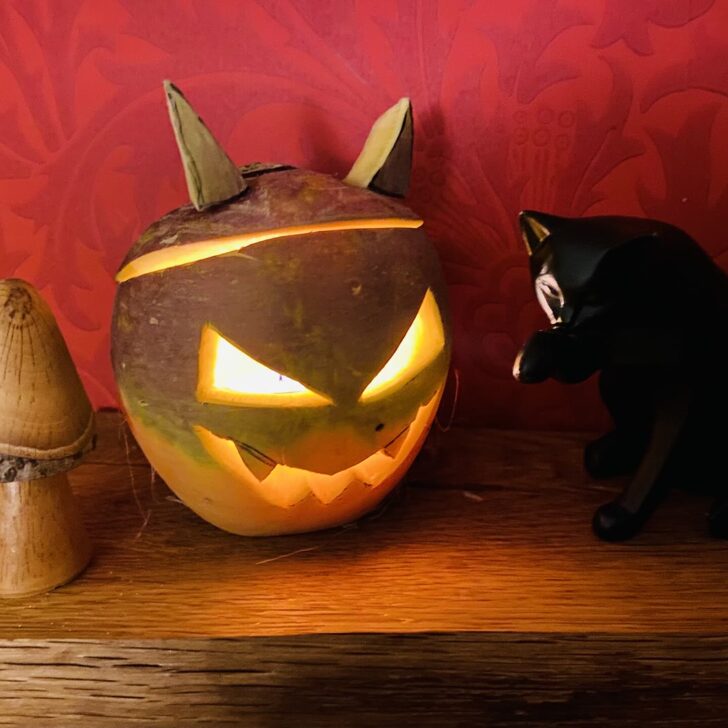 A carved turnip lantern with slanted glowing eyes, a jagged grin and pointed ears, lit from inside, beside a small wooden toadstool and a black cat ornament