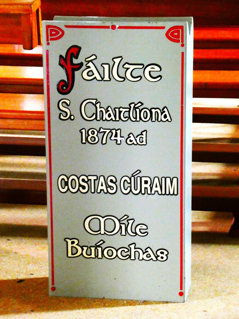 Painted church sign in Irish with a thin red border, reading F&aacute;ilte, S. Chaitl&iacute;ona 1874 ad, Costas C&uacute;raim and M&iacute;le Bu&iacute;ochas, standing in front of wooden pews