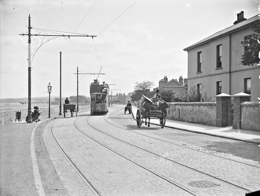 Black and white photograph of an open-top double-decker tram on a cobbled seafront road at Sandymount, Dublin, with horse-drawn carts, tall tram poles, a woman on a seat by the sea wall and a large two-storey house behind a garden wall