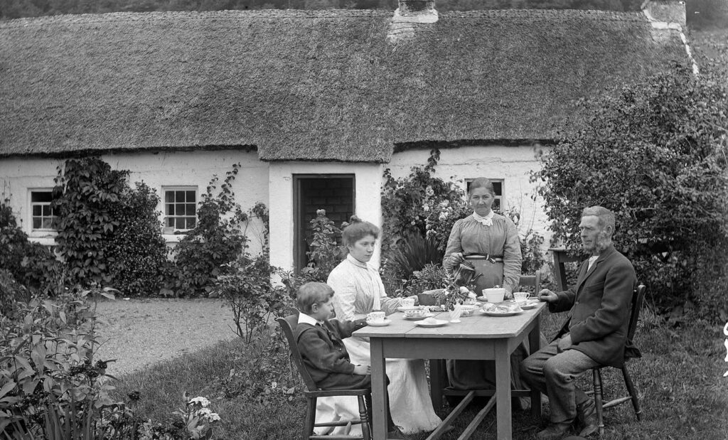 Black and white photograph from about 1900 of a family having tea at an outdoor table in front of a whitewashed thatched cottage at Glencar, as an older woman standing at the table pours for a young woman in a white dress, a small boy and an older bearded man