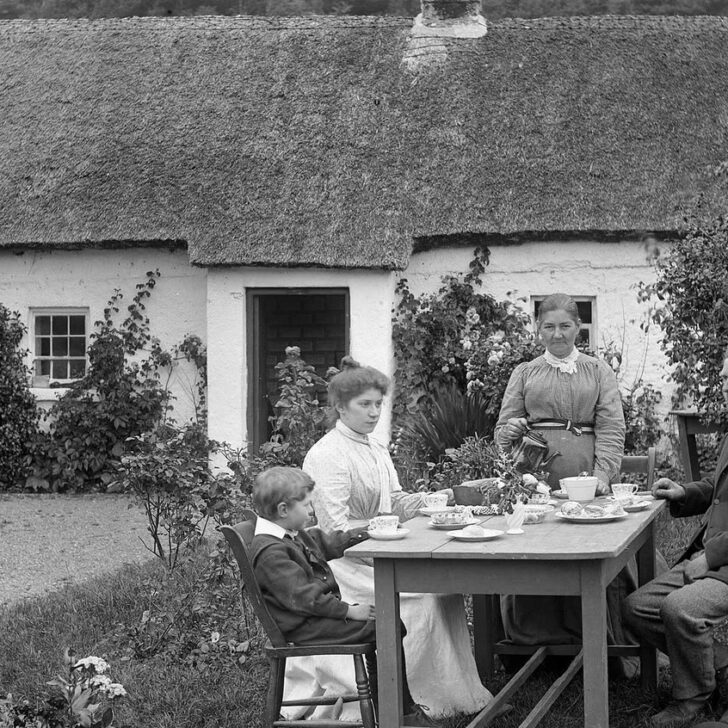 Black and white photograph from about 1900 of a family having tea at an outdoor table in front of a whitewashed thatched cottage at Glencar, as an older woman standing at the table pours for a young woman in a white dress, a small boy and an older bearded man