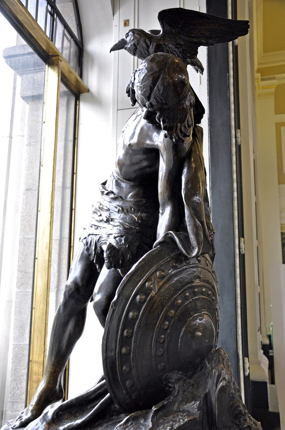 Dark bronze statue of the dying Cú Chulainn slumped forward, his round studded shield at his knee and a large bird with spread wings perched on his shoulder, beside a tall window in the GPO