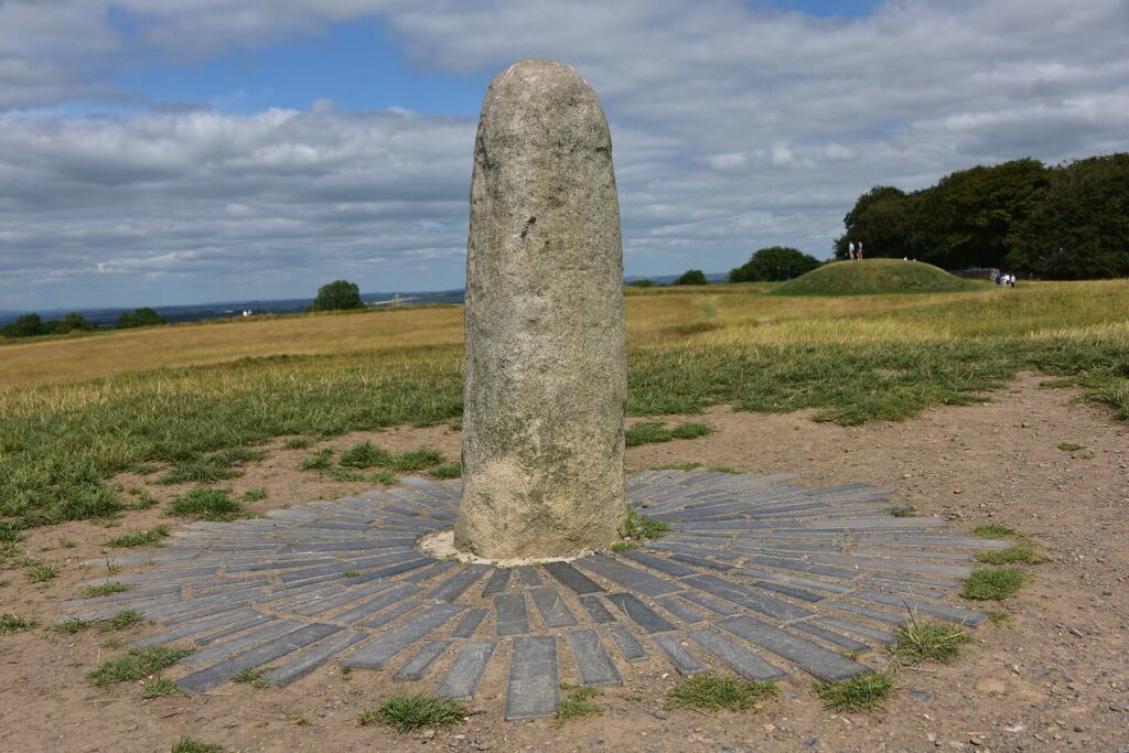 The Lia F&aacute;il on the Hill of Tara: a tall, rounded grey standing stone set in a ring of slate paving on a grassy hilltop, with a low earthwork mound and trees behind under a cloudy sky