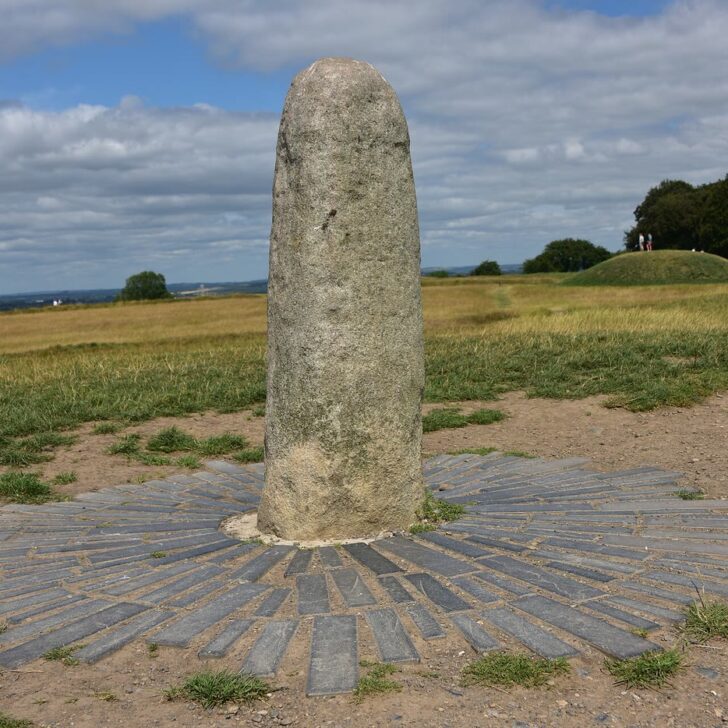 The Lia Fáil on the Hill of Tara: a tall, rounded grey standing stone set in a ring of slate paving on a grassy hilltop, with a low earthwork mound and trees behind under a cloudy sky