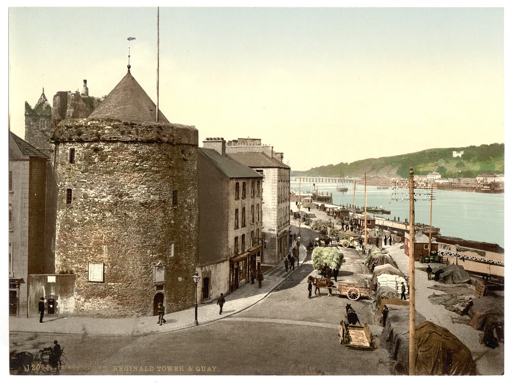 A colour-tinted photograph from about 1900 of Reginald's Tower, a round stone tower with a conical roof, at the corner of a street running along Waterford's busy quay beside the River Suir, with horse carts and small figures