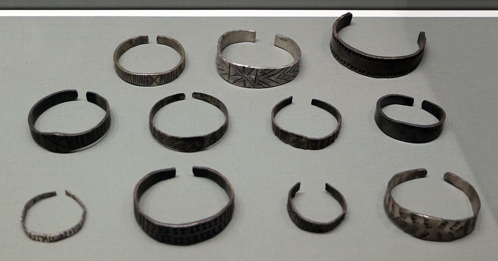 Plain and stamped silver arm-rings of different sizes, each open at one side, laid out in rows in a museum case