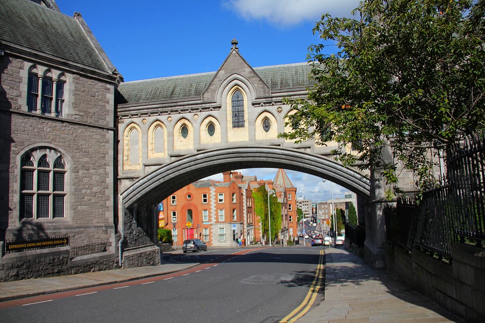 A Gothic stone bridge with arched windows spanning a Dublin street between Christ Church Cathedral and the Synod Hall, with red-brick buildings visible through the arch under a blue sky
