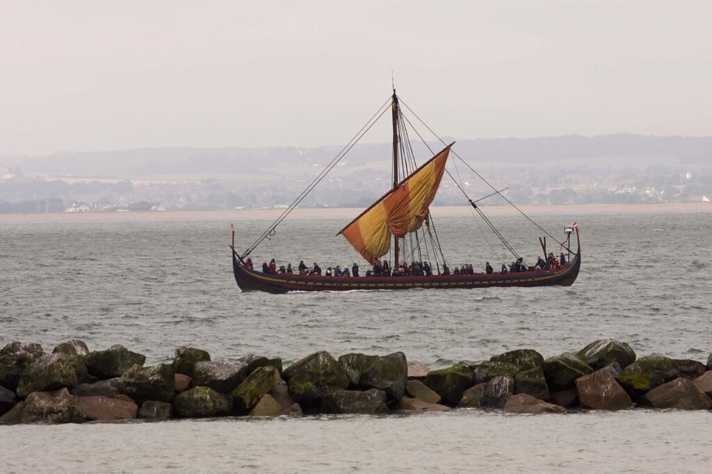A reconstructed Viking longship with a red and yellow striped sail being lowered and a row of crew along its side, on grey sea beyond a line of rocks, with a hazy coastline behind