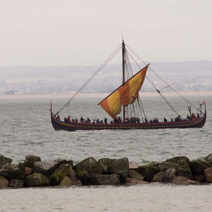 A reconstructed Viking longship with a red and yellow striped sail being lowered and a row of crew along its side, on grey sea beyond a line of rocks, with a hazy coastline behind