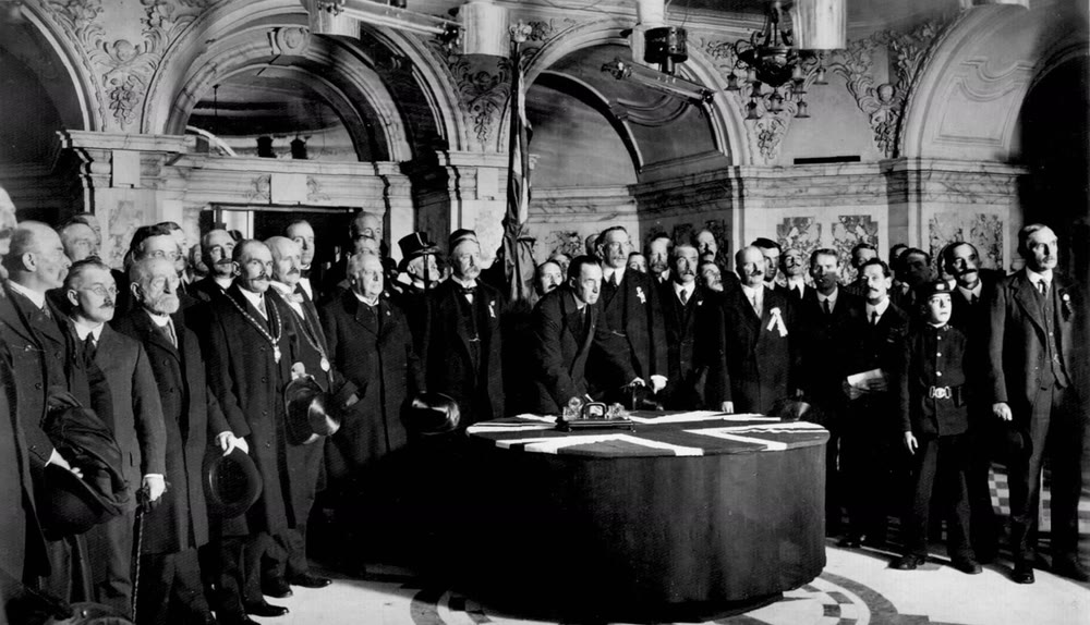 Black and white photo of Sir Edward Carson leaning over a round table draped with a Union flag to sign the Ulster Covenant, surrounded by dozens of men in dark suits in the ornate hall of Belfast City Hall