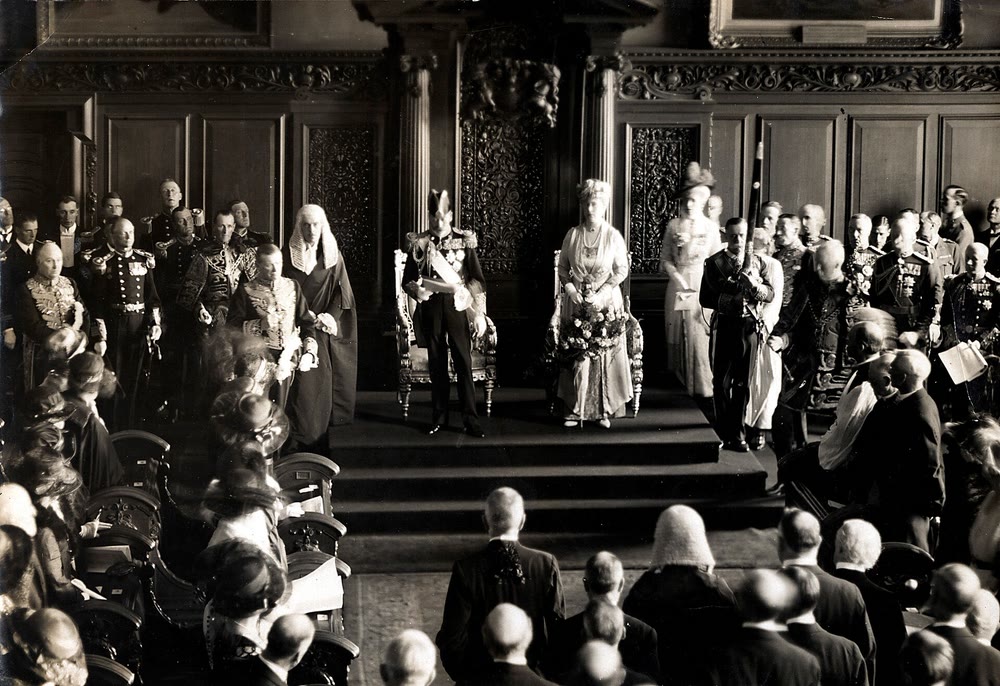 Sepia photo of King George V in uniform and Queen Mary standing on a dais in front of carved wooden panelling, with officials in robes, wigs and uniforms on either side and rows of seated guests in the foreground