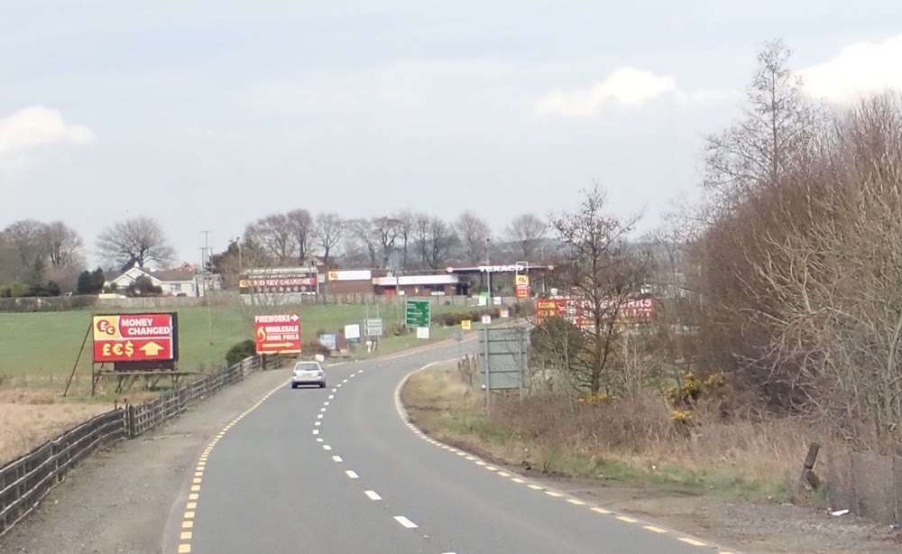 A quiet two-lane country road curving north into Northern Ireland, with a roadside sign offering to change money in pounds, euros and dollars and a petrol station in the distance