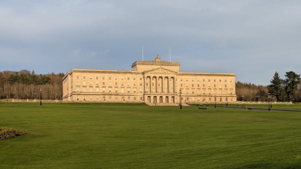 Parliament Buildings at Stormont, a long pale neoclassical building with a columned portico, at the top of a wide green lawn with woods behind