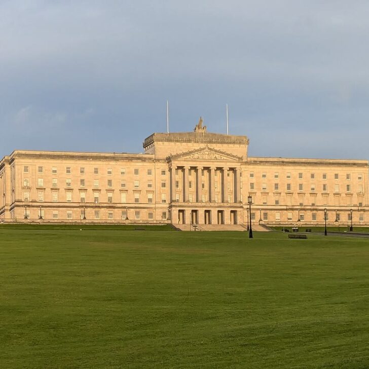 Parliament Buildings at Stormont, a long pale neoclassical building with a columned portico, at the top of a wide green lawn with woods behind